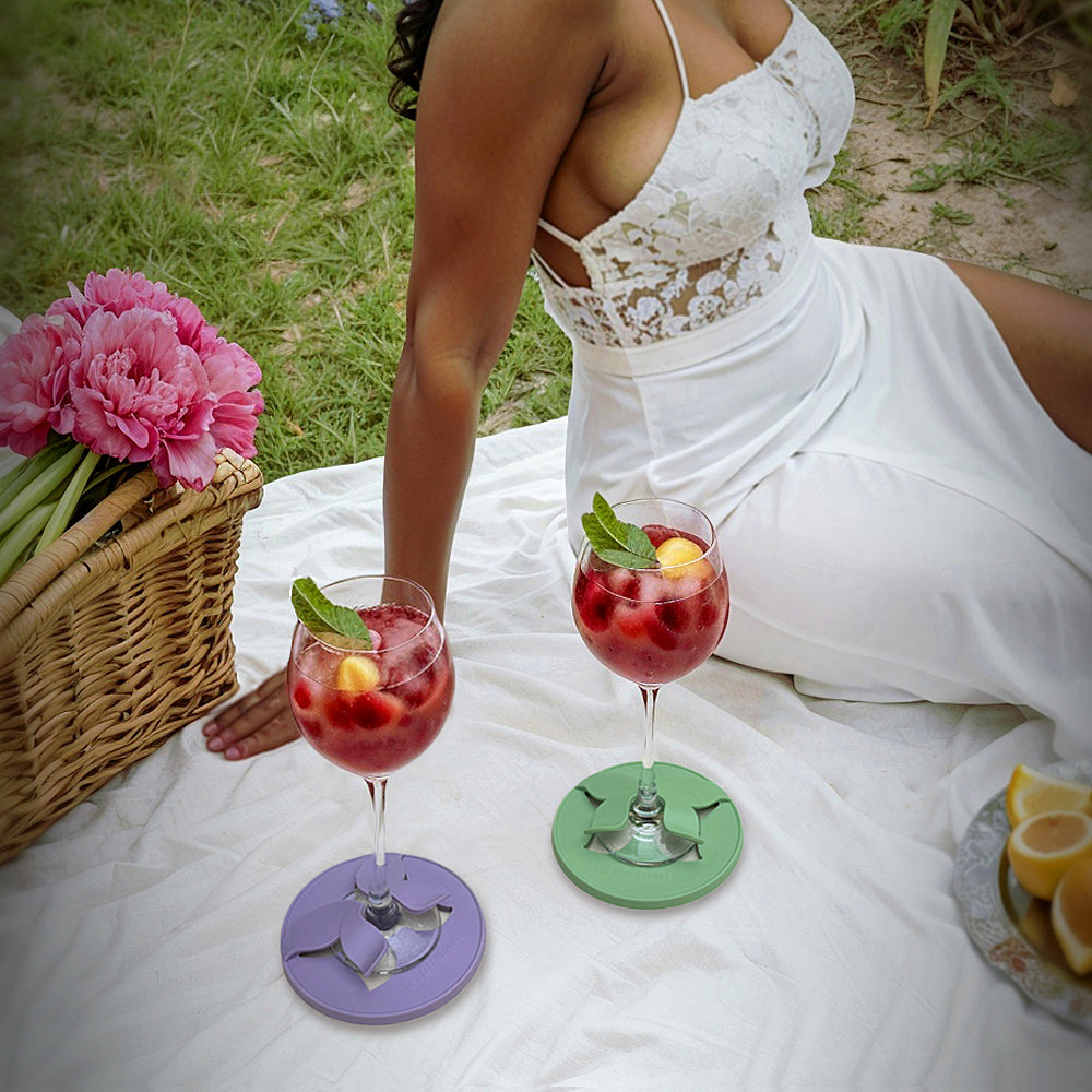 Wine glasses holding steady on a picnic blanket, each nestled in colorful Glass Slippers for stability, while a joyful African woman enjoys her cocktail against a beautiful outdoor backdrop